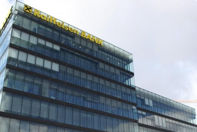 A contemporary glass office building prominently displays the Raiffeisen BANK name at the top. The structure features multiple stories with reflective windows, set against a partly cloudy sky. The yellow and black signage is clearly visible, indicating the financial institution's presence.