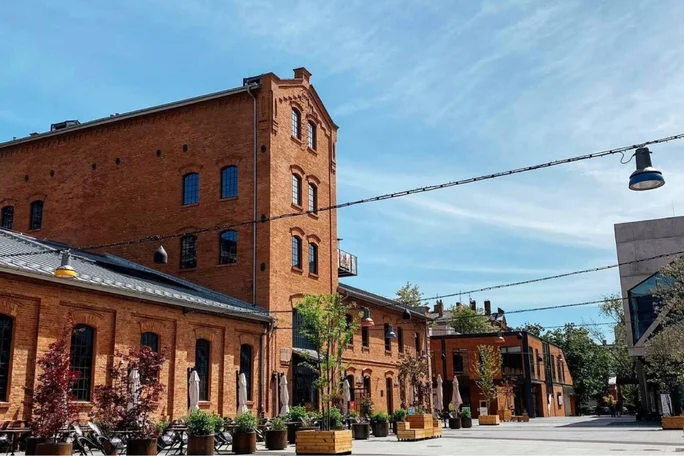 A large, multi-story brick building stands prominently in an open urban plaza under a clear blue sky. The setting features outdoor seating areas with umbrellas, potted plants, and string lights overhead, suggesting a relaxed and inviting atmosphere. The architecture highlights industrial heritage with arched windows and a mix of modern and historic elements. No visible text or numbers are present in the image.