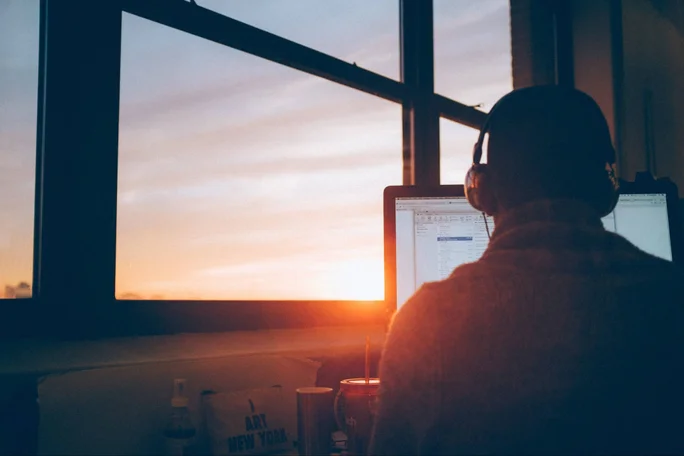 A person at a desk wearing headphones, gazing out the window at a beautiful sunset.